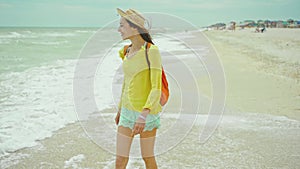 Happy carefree woman in hat walks alons sea water at sandy beach, enjoying find, sea waves and cloudy sky background. go