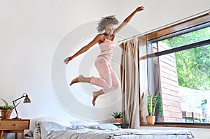 Happy carefree young African American woman wearing pajamas jumping on bed.