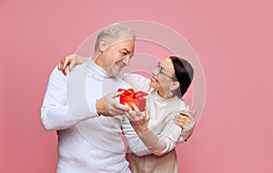 Happy, caring, loving senior couple, man and woman celebrating holiday, presenting gifts against pink studio
