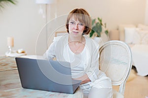 Happy casual beautiful woman working on a laptop sitting on the chair in the stylish white living room