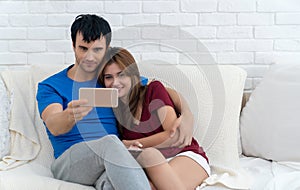 Happy casual couple taking themself a selfie or watching some digital content on smartphone sitting on white sofa in