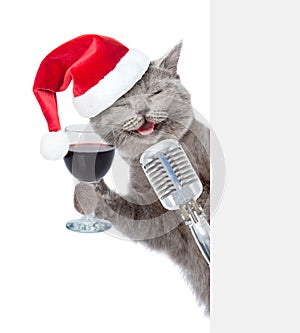 Happy cat with wineglass and retro microphone in red christmas hat peeking from behind empty board. isolated on white background