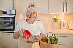 Happy Caucasian 60s woman sort vegetables from eco package in minimalist kitchen. Online buying food and grocery