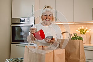 Happy Caucasian 60s woman sort vegetables from eco package in minimalist kitchen. Online buying food and grocery