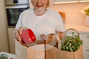 Happy Caucasian 60s woman sort vegetables from eco package in minimalist kitchen. Online buying food and grocery