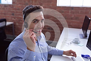Happy caucasian businessman wearing phone headset sitting at desk using computer smiling to camera