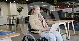 Happy caucasian casual businesswoman in wheelchair using laptop in office, in slow motion