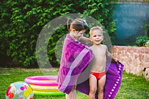 Happy Caucasian children play, wrapped in a large summer beach towel in the heat after swimming in a round home inflatable pool.