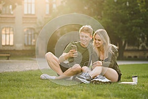 Happy caucasian college students using smartphone on campus lawn. Cheerful couple of students watching video on the phone, using