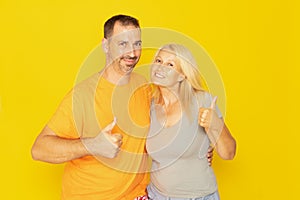 Happy caucasian couple in their 40s showing thumbs up and looking at camera isolated on yellow background.