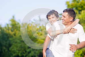 Happy Caucasian Family of Father and Son Piggybacking Outdoors. Against Nature Green Forest.