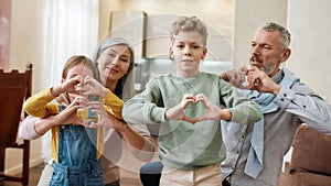 Happy caucasian family, grandparents and grandchildren making heart sign with hands and smiling at camera while sitting