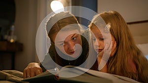 Happy Caucasian family loving caring mother reading book to little kid daughter using flashlight in bedroom lying on bed