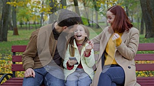 Happy Caucasian family mother father daughter sitting at bench in autumn park using mobile phone together parents with