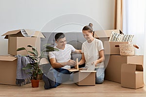 Happy Caucasian family moved in new apartment, sitting on the floor among cardboard boxes, couple in love rent a house together,