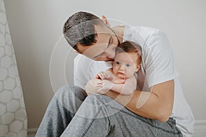 Happy Caucasian father holding newborn baby on laps knees. Man parent embracing kissing child daughter son. Authentic lifestyle