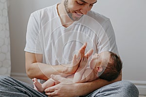 Happy Caucasian father holding newborn baby on laps knees. Man parent gives high five to child daughter son. Authentic lifestyle