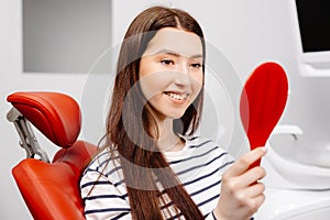 Happy caucasian female patient looking at mirror after dental treatment in clinic. Cheerful woman sitting in chair in dental