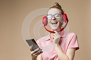 Happy Caucasian Girl In Casual Clothing Posing With Wireless Headphones And Cellphone While Laughing Against Beige