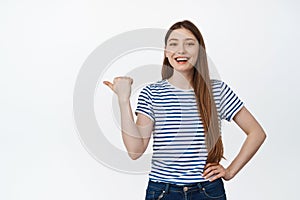 Happy caucasian girl laughing, smiling and casually pointing finger left at empty space, shopping sale banner, standing