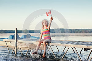 Happy Caucasian girl sitting on dock pier by lake and waving Canadian flag. Smiling child holding Canada flag sitting by water.