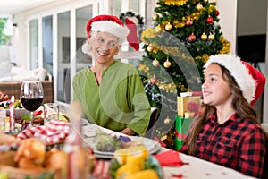 Happy caucasian grandmother and granddaughter wearing santa hats talking at christmas table