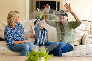 Happy caucasian grandmother and grandson watching grandfather wearing vr headset on couch at home