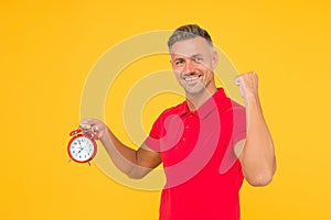 Happy caucasian guy flex strong arm holding alarm clock yellow background, punctuality