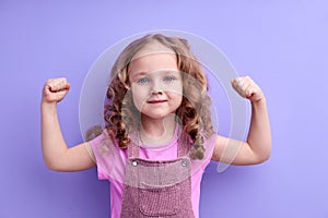 Happy caucasian little child girl showing arm muscles looking at camera isolated on purple