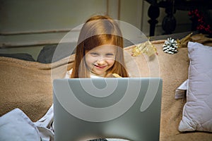 Happy caucasian little girl during video call or messaging with Santa using laptop and home devices, looks delighted and