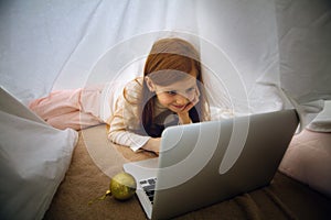 Happy caucasian little girl during video call or messaging with Santa using laptop and home devices, looks delighted and