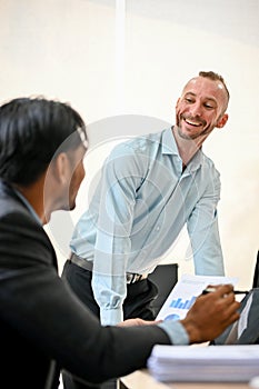 Happy caucasian male boss enjoys talking with his Asian male coworker during the meeting