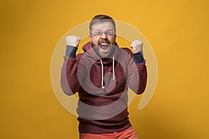 Happy Caucasian man makes a winner gesture, he energetically raises fists up and shouts in joy. Yellow background.