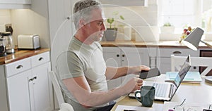 Happy caucasian man sitting at table in kitchen, using laptop and talking on smartphone