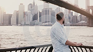 Happy Caucasian man stands near river embankment fence at Brooklyn Bridge scenery, reflecting and enjoying New York view