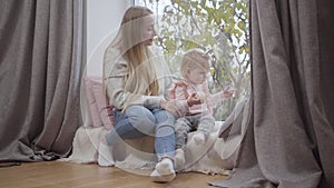 Happy Caucasian mother and daughter sitting at the windowsill and looking out through the window. Cute blond baby girl