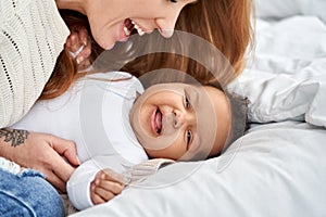 Happy caucasian mother having fun with cute little baby daughter on bed.