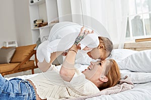 Happy caucasian mother having fun lifting cute little baby daughter in bed.