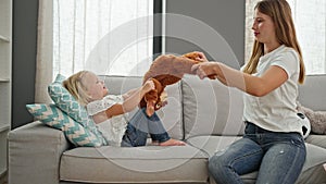 Happy caucasian mother and little daughter comfortably relaxing on the sofa, playing together with a monkey doll in their cozy