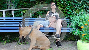 Happy caucasian mother and son with dog on a bench in the park. Urban green space. Bright sunny summer day