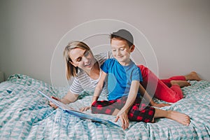 Happy Caucasian mother and son family reading book in bedroom. Mom and boy child at home spending time together. Parent talking