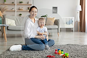 Happy caucasian mother and toddler son smiling at camera, playing together, sitting on floor carpet in living room