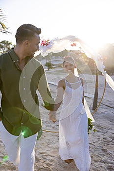 Happy caucasian newlywed couple holding hands and walking at beach at wedding ceremony at sunset