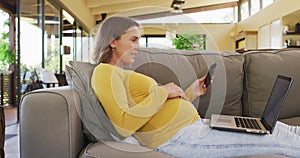 Happy caucasian pregnant woman sitting in armchair and using smartphone and laptop