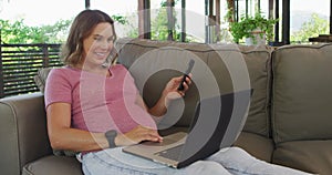 Happy caucasian pregnant woman sitting on sofa and using smartphone and laptop