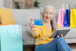 Happy caucasian retired gray-haired woman sits on floor with tablet and a lot of shopping bags, shows credit card
