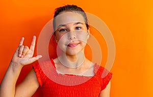 Happy caucasian schoolgirl doing sign language with hand over orange background