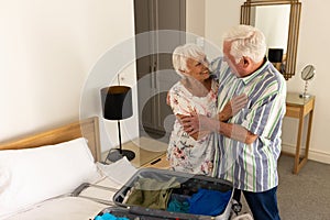 Happy caucasian senior couple packing suitcase and embracing in sunny bedroom at home