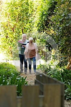 Happy caucasian senior couple walking with flowers and watering can in sunny garden