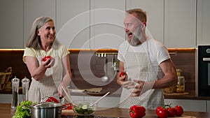 Happy Caucasian senior old couple having fun cooking at home funny man fooling around juggling tomatoes smiling woman